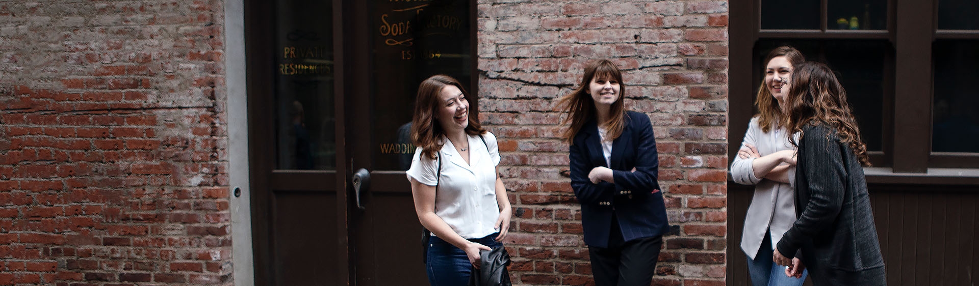 Group shot showing 4 women chatting and laughing together, leaning against a red brick building