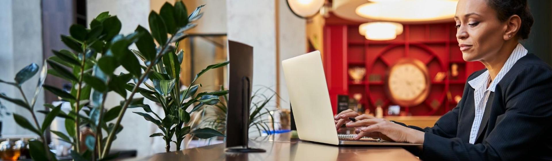 Woman in a blazer working on a laptop at a desk. She is in front of a red background and there are plants in the left foreground. 