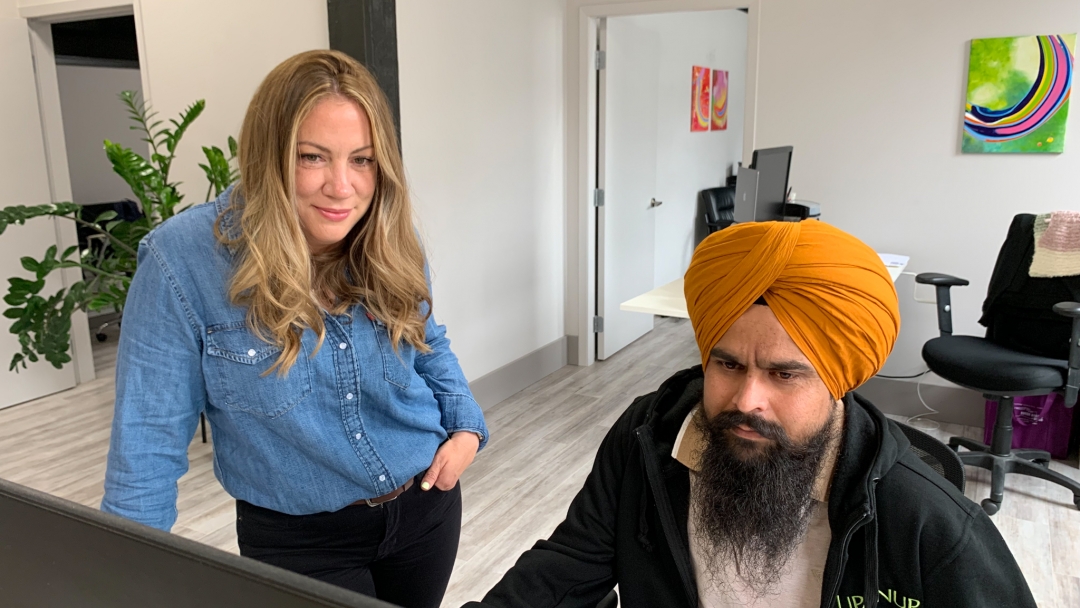 Woman standing beside a man sitting in a chair looking at a computer. 