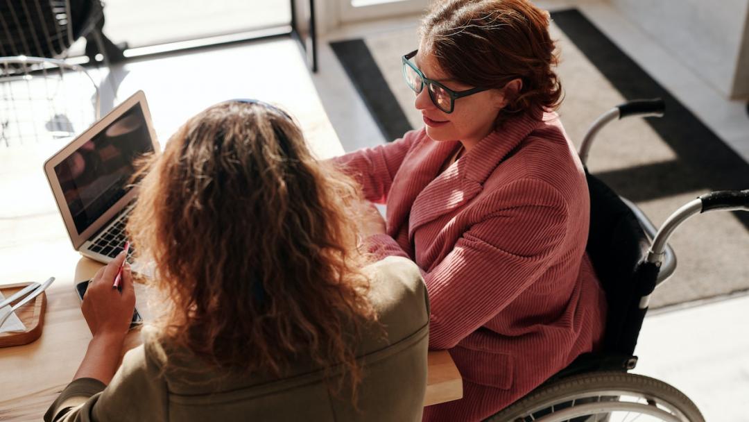 Two women sitting in front of a laptop, one is in a wheelchair. 