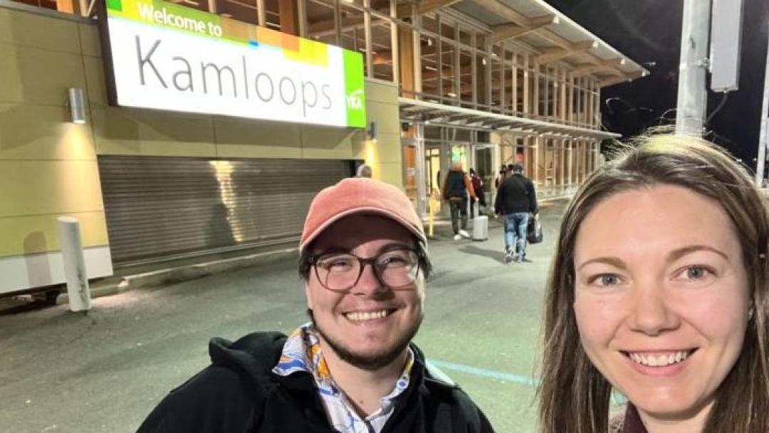 Two people taking a selfie smiling in front of the welcome to kamloops building sign.