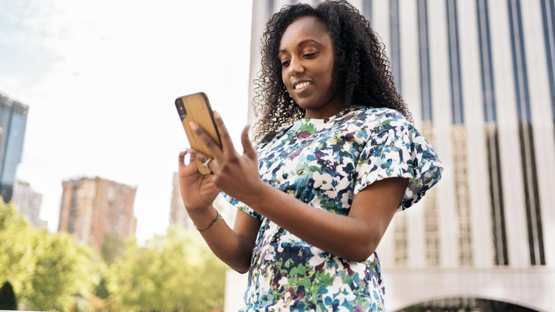 Looking up to a young black woman holding a phone with both hands. There's a skyscraper in the background on the right, and trees with smaller buildings in the left background.