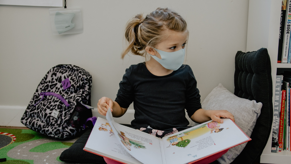 Young girl reading a book with a mask on. 