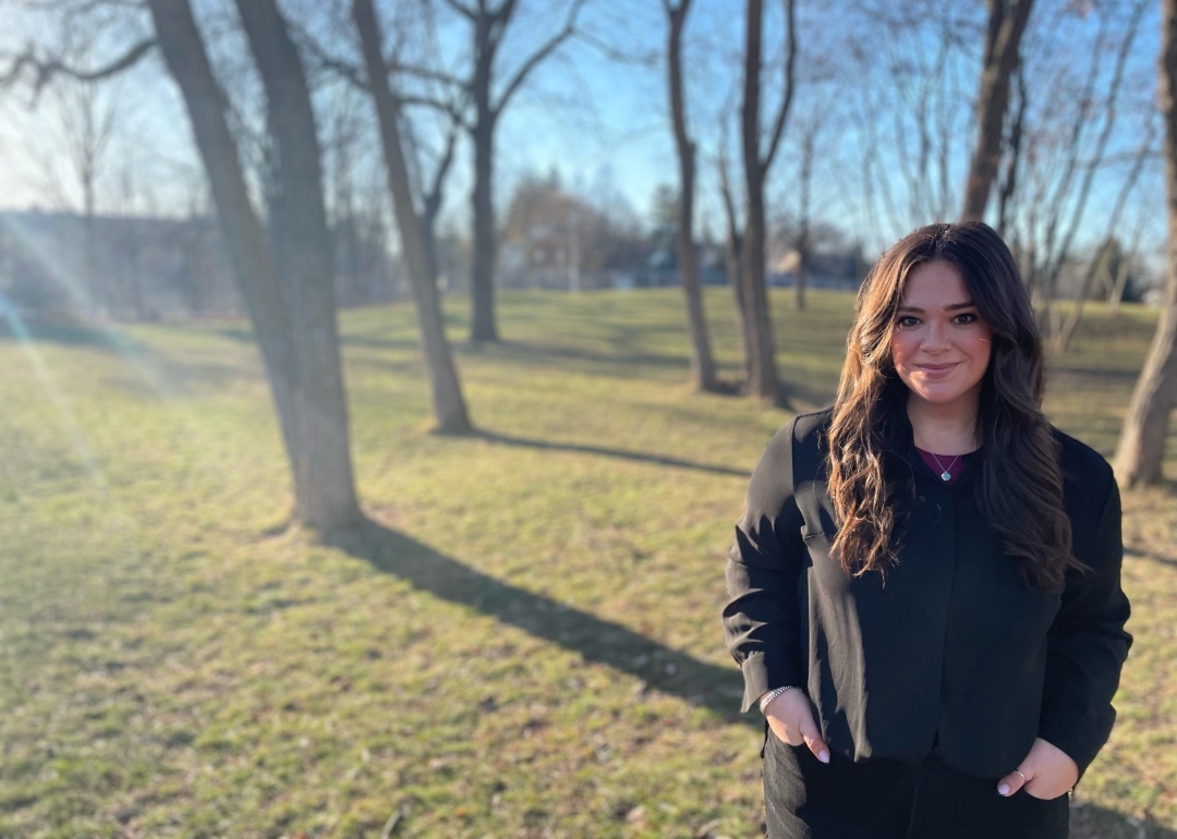 Woman with brown hair standing outside by the trees. 