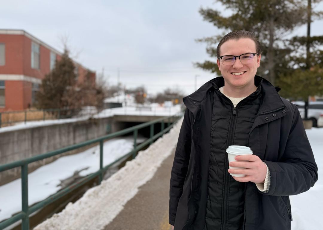 A person holding a coffee smiling at the camera with a building, pathway and snow behind them.