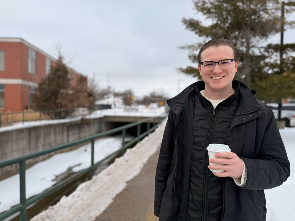 A person holding a coffee smiling at the camera with a building, pathway and snow behind them.